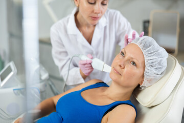Young female cosmetologist performs hardware facial exfoliating procedure to adult female patient