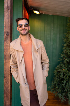 Handsome man in beige stylish trench in front of wooden house. Elegant male wearing salmon-colored sweater and pale brown trench coat standing outdoors. Fashionable guy in sunglasses winter holidays