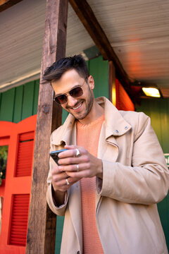 Man in beige trench coat using phone. Handsome guy in trench and sunglasses smiling at his smartphone outdoors. Fashionable guy leans on wooden pillar while chatting with mobile phone