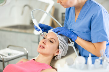 Adult woman cosmetologist performs hardware facial procedure to young woman patient