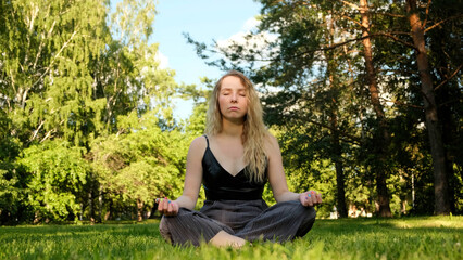 Young beautiful relaxed blonde woman sitting on green grass and meditating. Concept. Unity with nature, meditation in a city park.