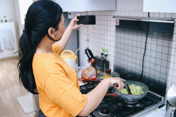 Young Asian woman taking selfie with smartphone while cooking in the kitchen - obrazy, fototapety, plakaty