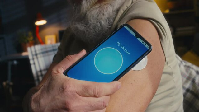 Close-up shot of anonymous senior man in sleeveless vest putting smartphone to blood glucose monitor patch on shoulder, measuring with mobile app and getting normal reading