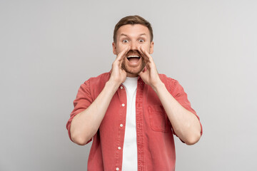 Amazed young man shouting with hands around mouth looking at camera on gray background. Positive...