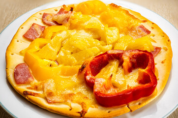 Closeup view of delicious homemade hawaiian pizza topping with pieces of pineapple, slice of bell chilli and ham in white ceramic plate on plastic table mat.