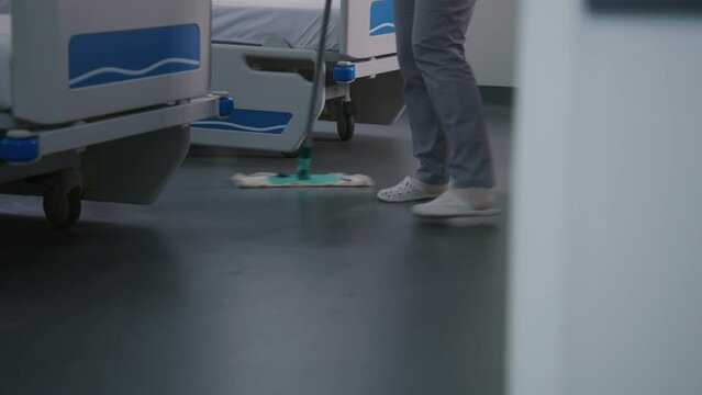 Close up of adult nurse mops floor near beds in bright hospital ward. Health workers take care of cleanliness in medical center. Cleaner prepares hospital room for new patients. Medical staff at work.