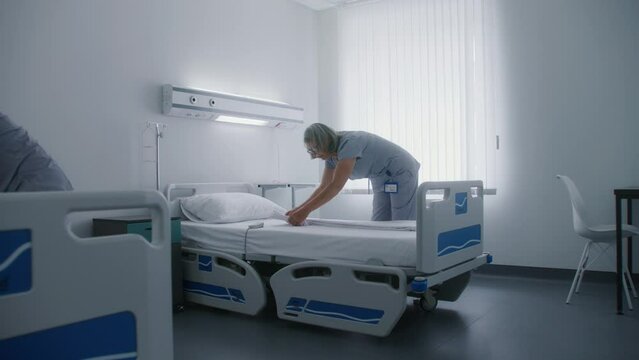 Mature cleaner with her adult colleague make beds in modern and bright hospital ward. Female health workers change bedclothes on beds after patients. Medical staff at work in modern medical center.