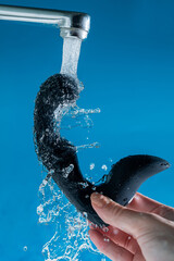 A woman washes a black prostate stimulator under running water on a blue background. Sex toy...