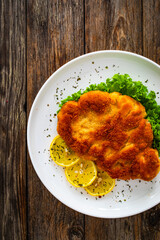 Crispy breaded seared chicken cutlet with lemon slices and fresh vegetables on wooden table
