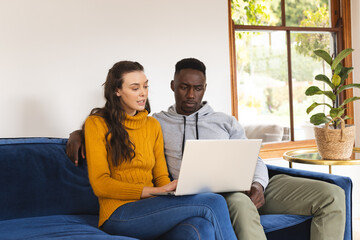 Diverse couple sitting on sofa using laptop at home