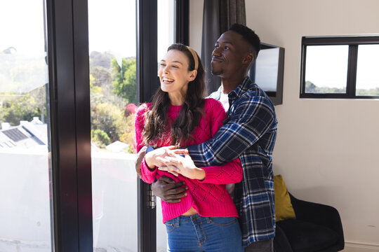 Happy diverse couple embracing and looking through window at home