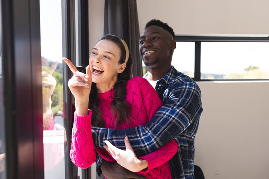 Happy diverse couple embracing and looking through window at home