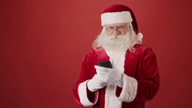 Medium shot of cheerful bearded senior man in Santa Claus costume taking selfie on smartphone, then typing merry Christmas message to someone, on red background. Template, copy space