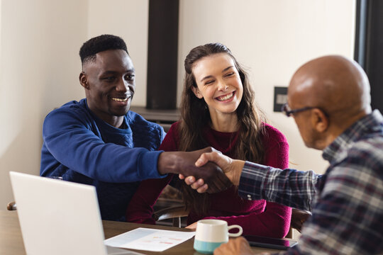 Diverse couple and biracial male financial advisor using laptop and discussing documents at home