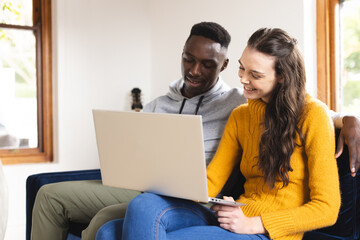 Happy diverse couple sitting on sofa using laptop at home