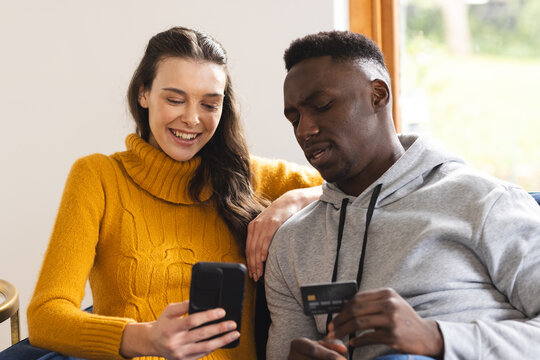 Happy diverse couple sitting on sofa using smartphone for online shopping at home