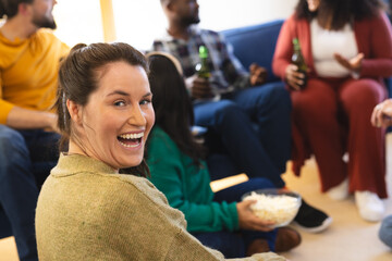 Happy diverse male and female friends talking, eating popcorn and drinking beer at home