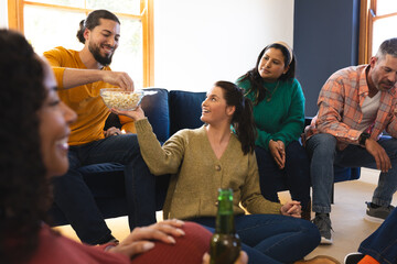Happy diverse male and female friends talking, eating popcorn and drinking beer at home