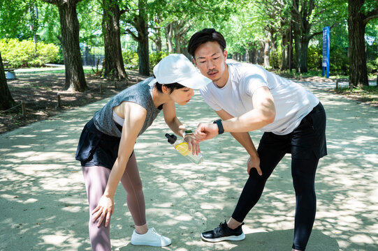 East Asian man and woman in sportswear check their smartwatches on a green park boulevard. Holding a water bottle. smiling and stretching.Healthy living. Physical and mental wellness.