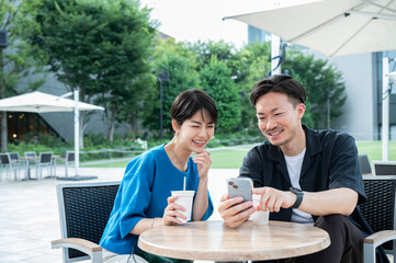 Beautiful woman smiling and talking to man. Park with lots of greenery. Chatting on a café terrace with coffee and smartphone in hand. Happy city life.