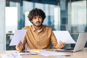 Fototapeta Portrait of young dissatisfied and disappointed financier, businessman behind paperwork looking at camera, negative checking documents and financial reports inside office, using laptop at work. obraz