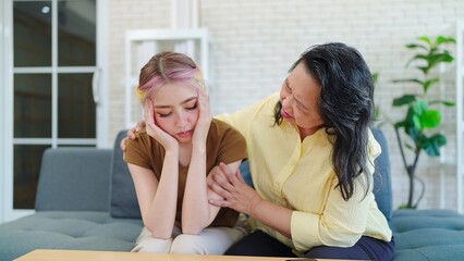 Asian eldery mother taking care young daughter having suffer from migraine or headache while sitting on sofa at home. Mother hugging unwell young daughter with headache. Healthy problem concept - obrazy, fototapety, plakaty