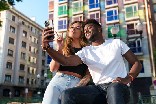 Two attractive, diverse models in urban casual attire share a joyful selfie moment against a backdrop of colorful city buildings.