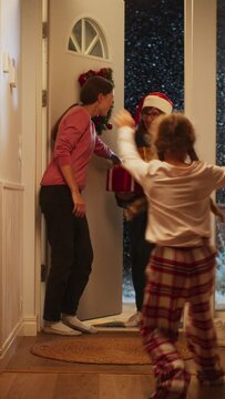Vertical Screen: Young Mother Meeting Grandparent by the Door in a Decorated Home with Garlands, Small Boy and Girl Running to Meet Their Grandmother with Christmas Gifts and Presents