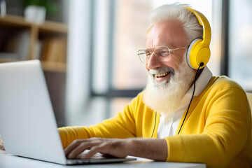 Happy old man with white hair and beard in yellow headphones and casual clothing calling video using his laptop. Online meeting. Video Call.
