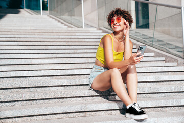 Obraz premium Young beautiful smiling hipster woman in trendy summer clothes. Carefree woman with curls hairstyle, in the street. Positive model holds mobile phone, looks at cellphone screen, uses smartphone apps