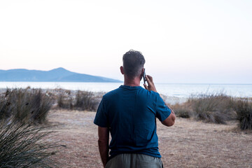 Back view of standing man using mobile phone making phone call in outdoor against a travel scenic destination background with ocean and island. People tourist and cellphone technology connection wifi