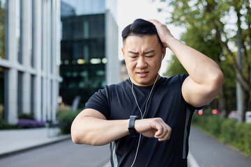 Upset young asian man standing in the middle of the street and looking at the smart watch, not happy with the result, holding his head in frustration