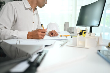 Focused architect male sitting at desk with architectural model and designing layout of buildings at workstation