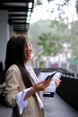 Portrait of cheerful millennial businesswoman holding mobile phone and laughing while standing near office building