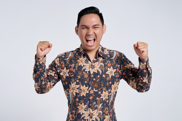 Excited young Asian teacher man wearing batik shirt celebrating success with raised fist isolated...