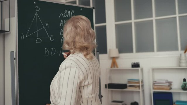 Woman geometry teacher writing formula on chalkboard during lesson, mathematics