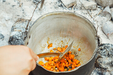 Hand of man holding skimmer and stirring carrot with spices, preparing for cooking meal in cauldron...