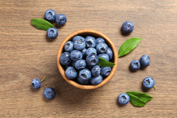 Bowl of tasty fresh blueberries with green leaves on wooden table, flat lay