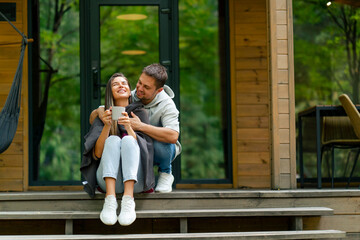 A thoughtful woman sits on the doorstep of a wooden house with cup of tea with her caring husband