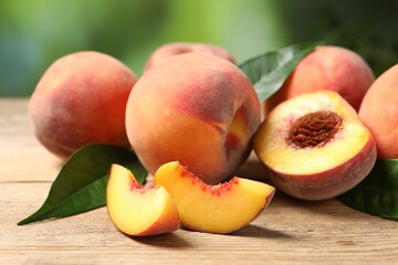 Cut and whole fresh ripe peaches with green leaves on wooden table against blurred background, closeup