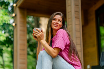 A thoughtful girl sits with a cup of tea on the terrace of her wooden cottage in the woods in...