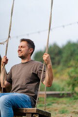 A young handsome man smiles and rides a large wooden swing in field in nature overlooking a lake