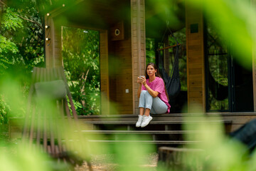 A thoughtful girl sits with a cup of tea on the terrace of her wooden cottage in the woods in...