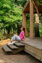 Fototapeta premium A thoughtful girl sits with a cup of tea on the terrace of her wooden cottage in the woods in morning