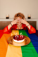 very happy woman covers her eyes in front of her surprise, cake with candles on top of lgtb tablecloth
