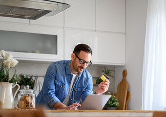Businessman with credit card shopping online over laptop on kitchen counter while working from home