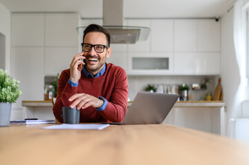 Cheerful businessman speaking over smart phone while sitting with laptop at desk in home office
