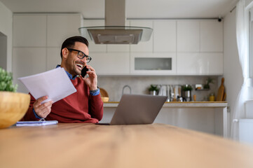 Male entrepreneur with spreadsheet having business call over smart phone while working at home