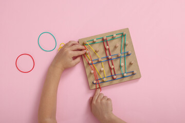 Motor skills development. Boy playing with geoboard and rubber bands at pink table, top view