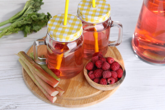 Mason jars of tasty rhubarb cocktail with raspberry and stalks on white wooden table